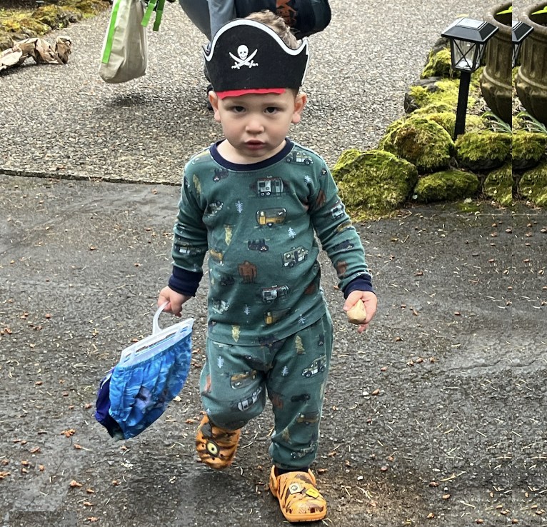 A toddler in patterned pajamas and a paper pirate hat walking down a driveway, carrying a bag