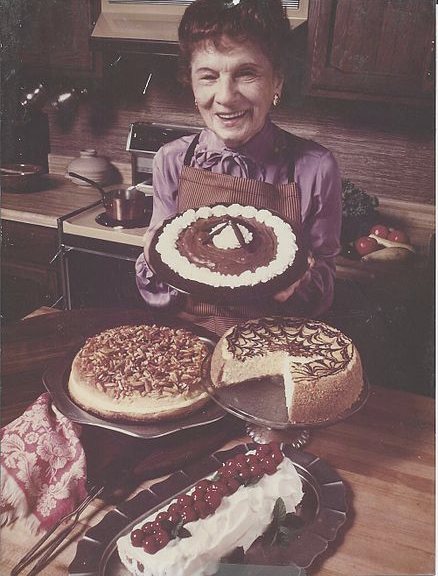 Rose Naftalin in her kitchen, presenting a chocolate cream pie with cheesecakes and pastries on the table